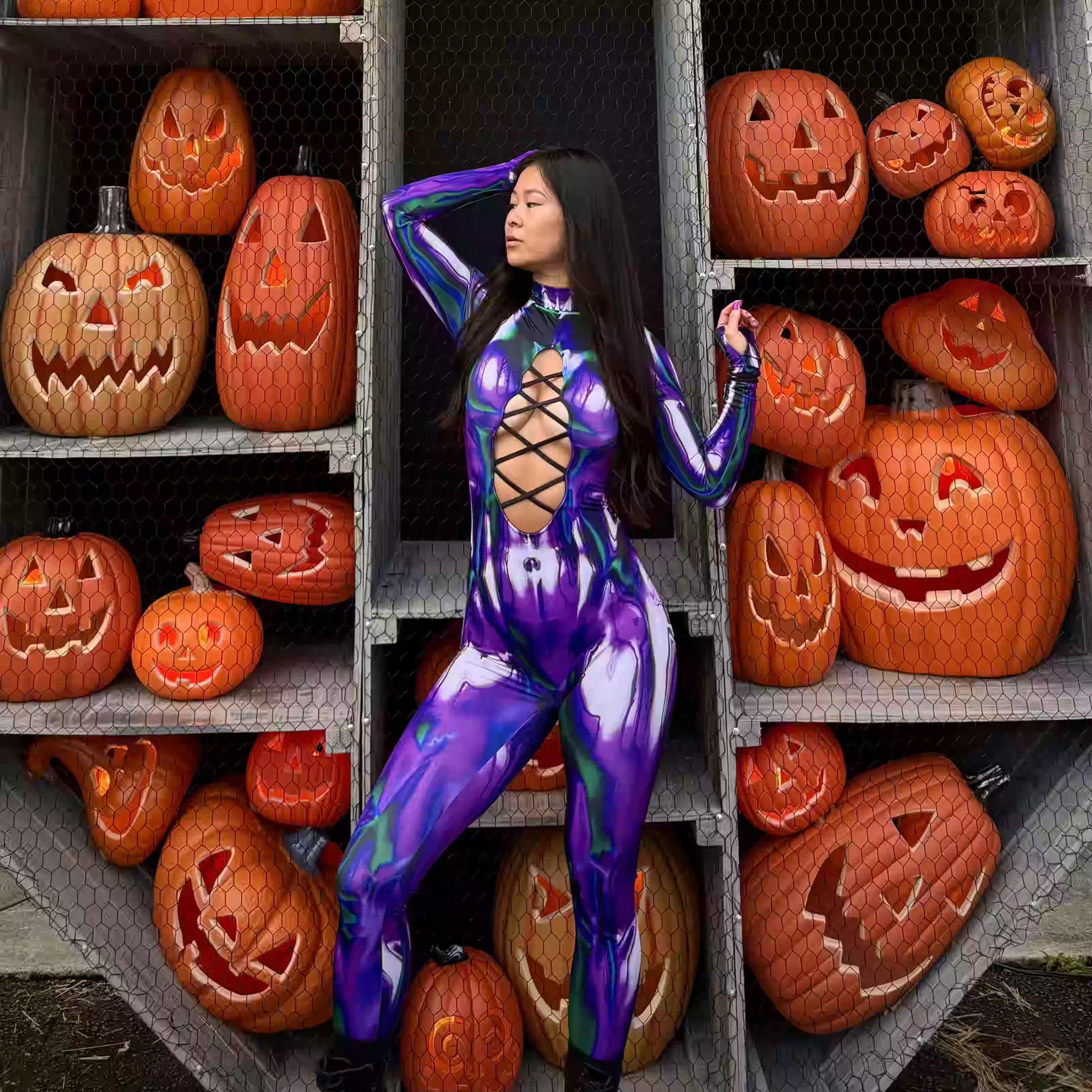 Woman wearing a purple and green long-sleeve cut-out Halloween catsuit, posing in front of carved jack-o'-lanterns, a bold rave outfit idea for BOO Seattle.