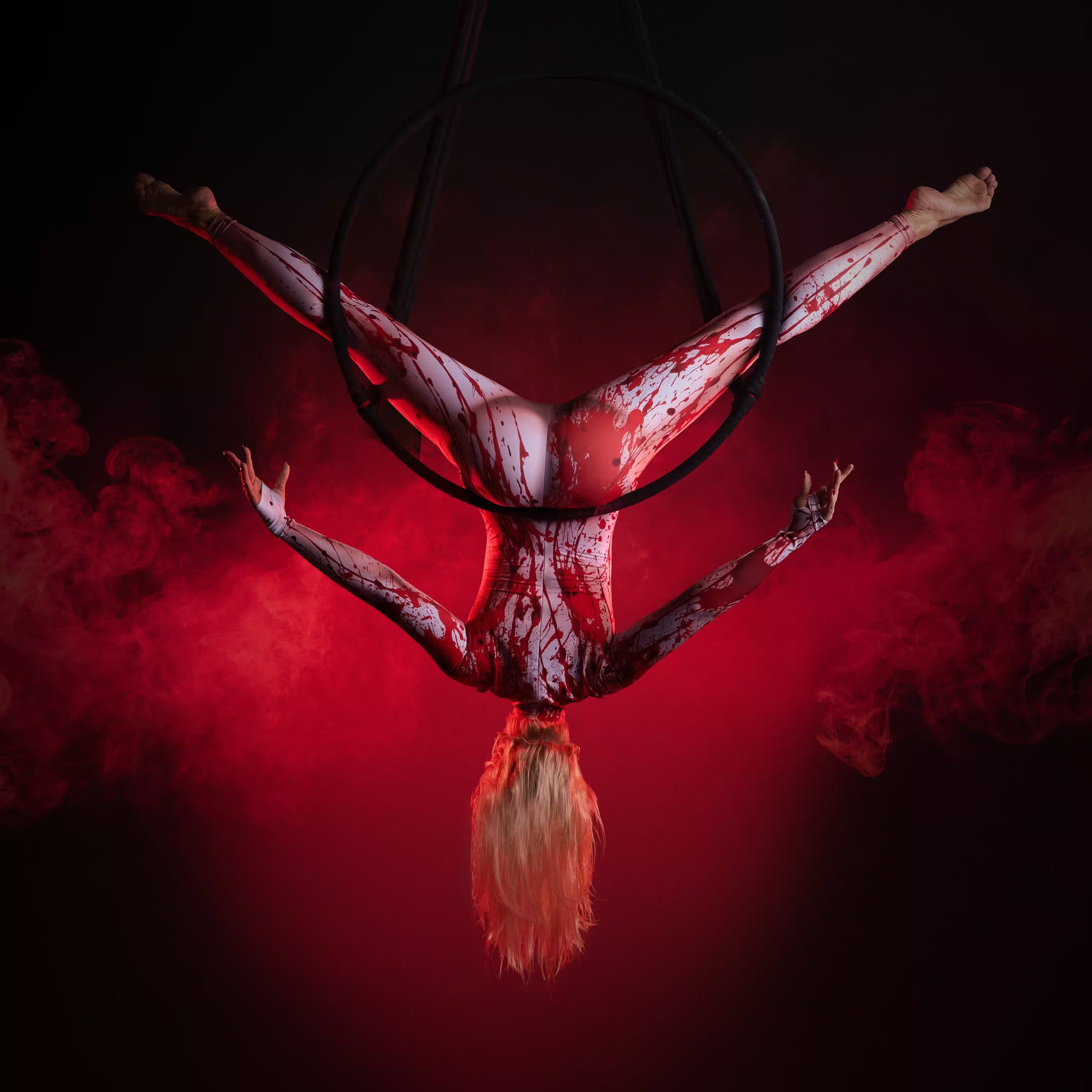 Blonde aerial performer wearing a white blood-splattered bodysuit hangs upside down from an aerial hoop surrounded by red smoke.