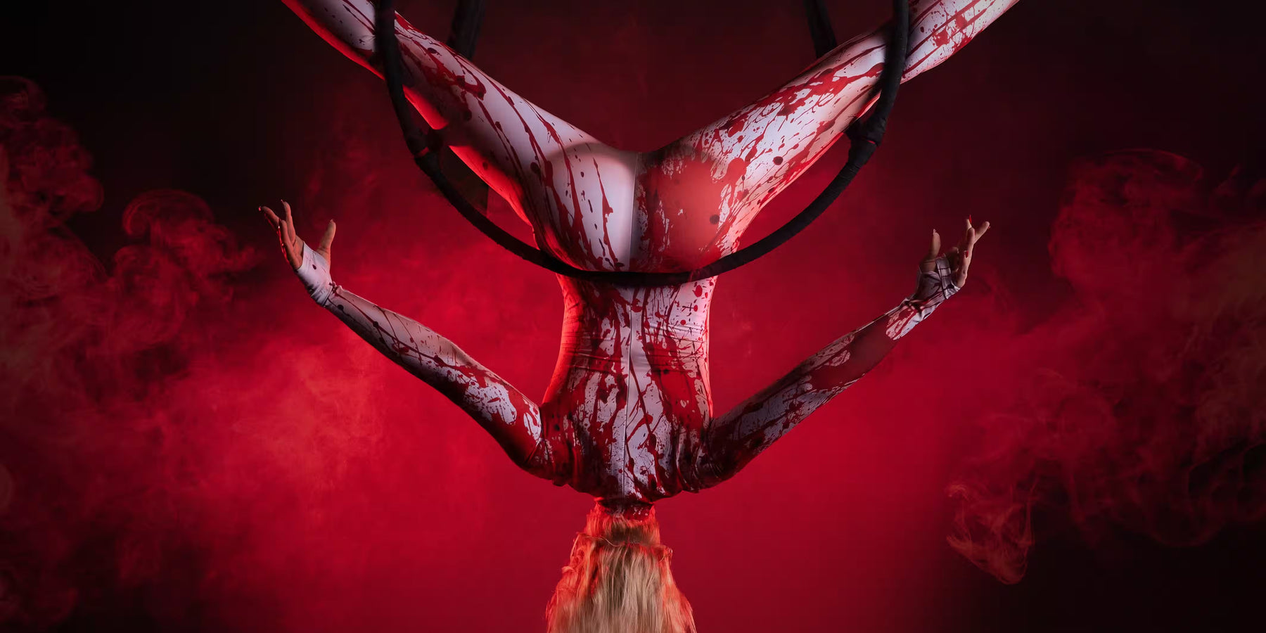 Blonde aerial performer wearing a white blood-splattered bodysuit hangs upside down from an aerial hoop surrounded by red smoke.