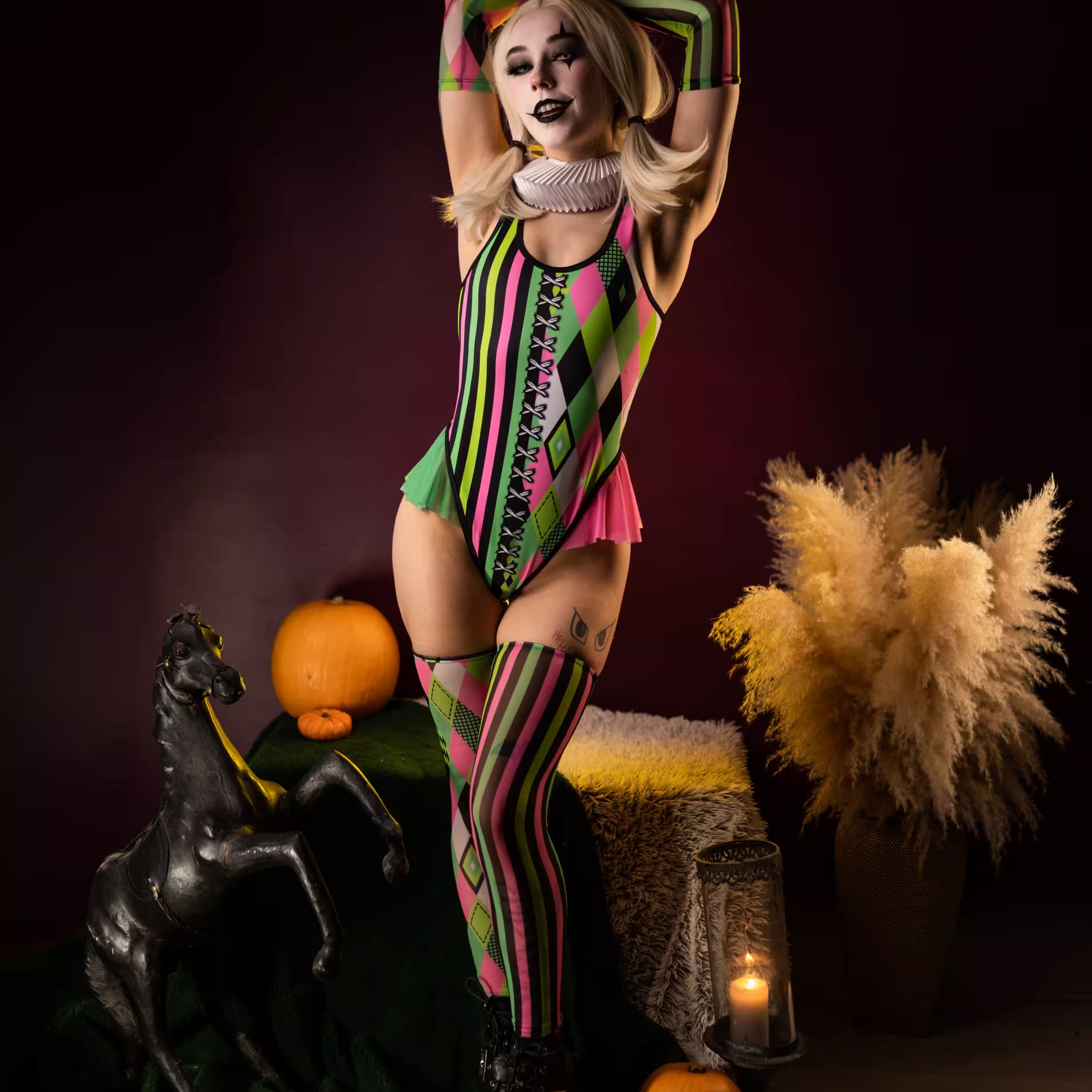 Woman wearing a colorful pink, green and black jester bodysuit with matching stockings and clown makeup, posing beside pumpkins and Halloween decorations.