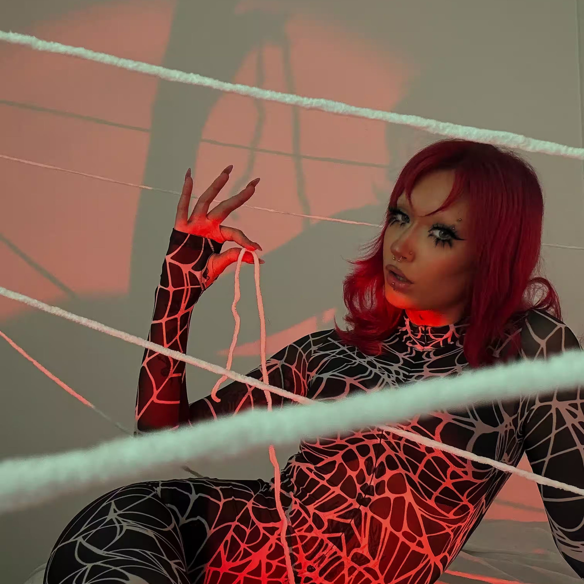 Woman with red hair wearing a black spiderweb-print bodysuit and dramatic eye makeup, posing among white webs under red lighting.