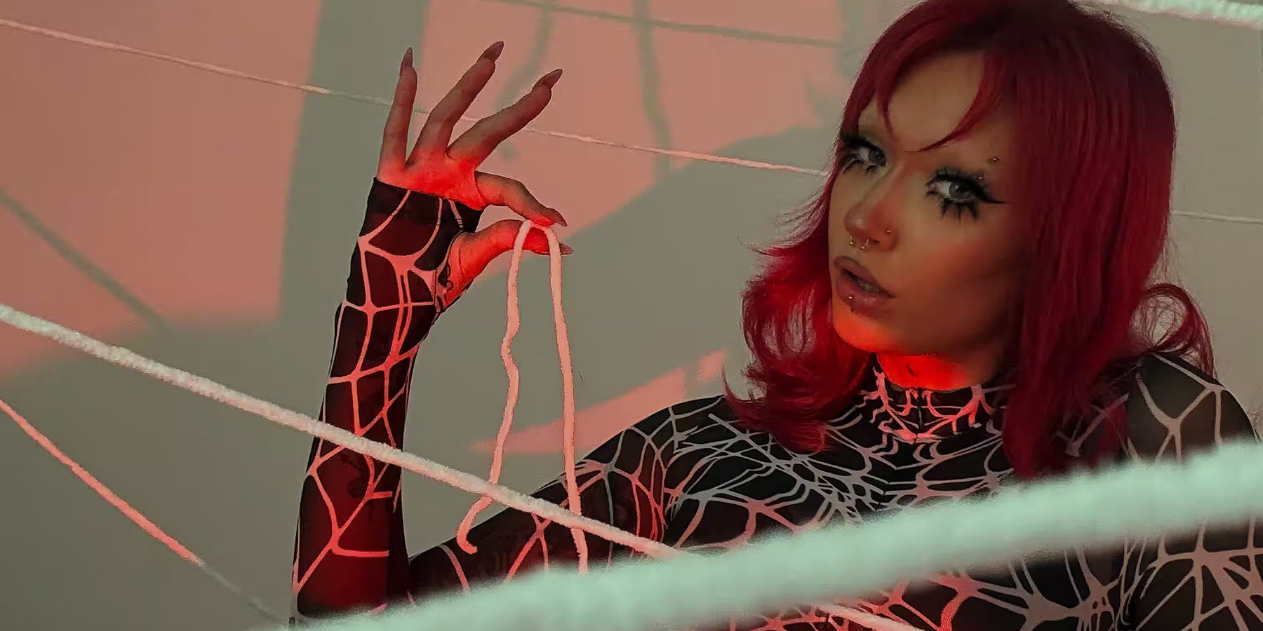 Woman with red hair wearing a black spiderweb-print bodysuit and dramatic eye makeup, posing among white webs under red lighting.