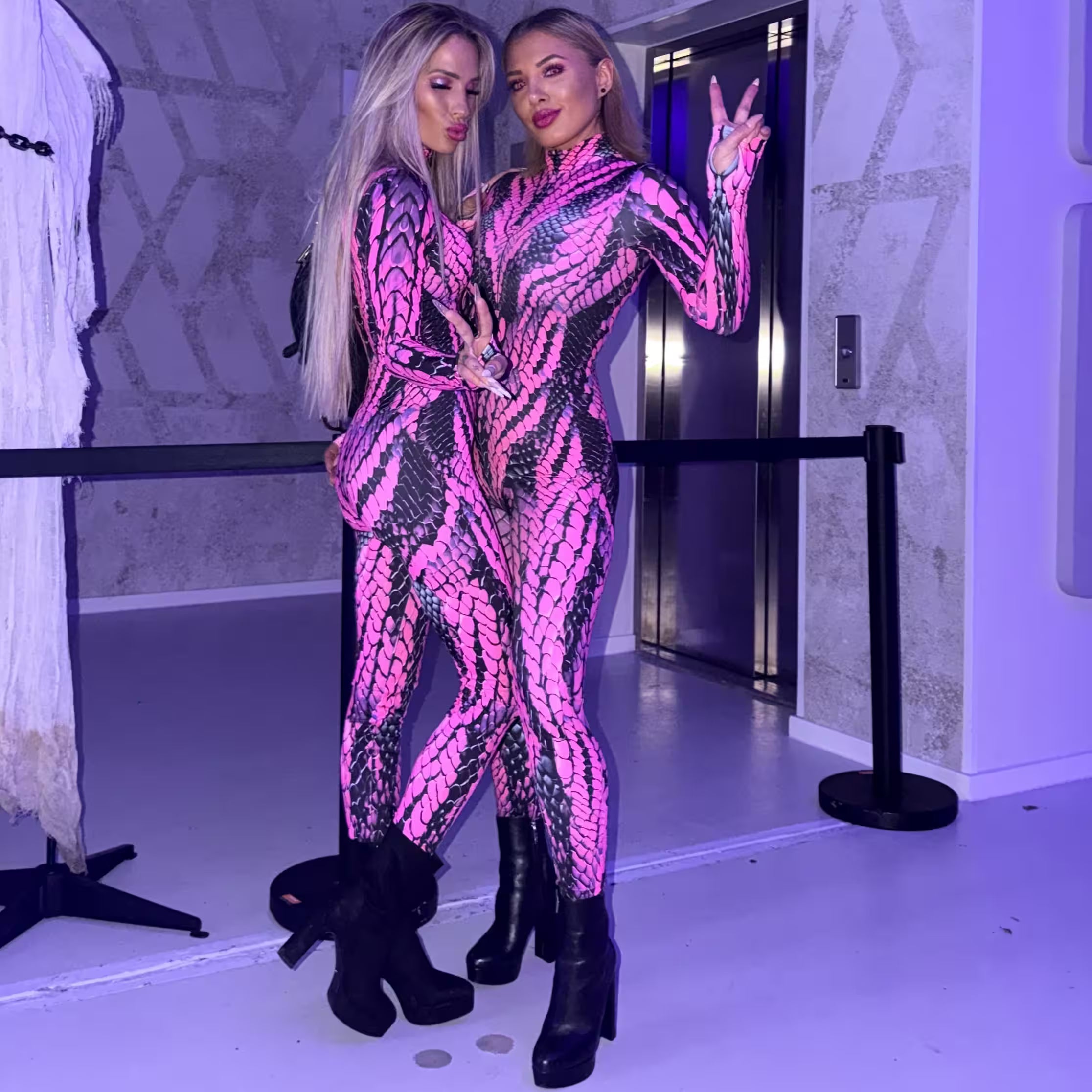 Two women wearing matching pink and black snakeskin catsuits with black platform boots, posing together in Halloween rave outfits for MÁS EDM Los Angeles.