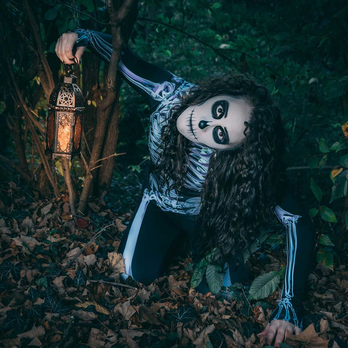 Woman wearing a black skeleton bodysuit and skull face makeup crouches in a dark forest while holding a glowing lantern.