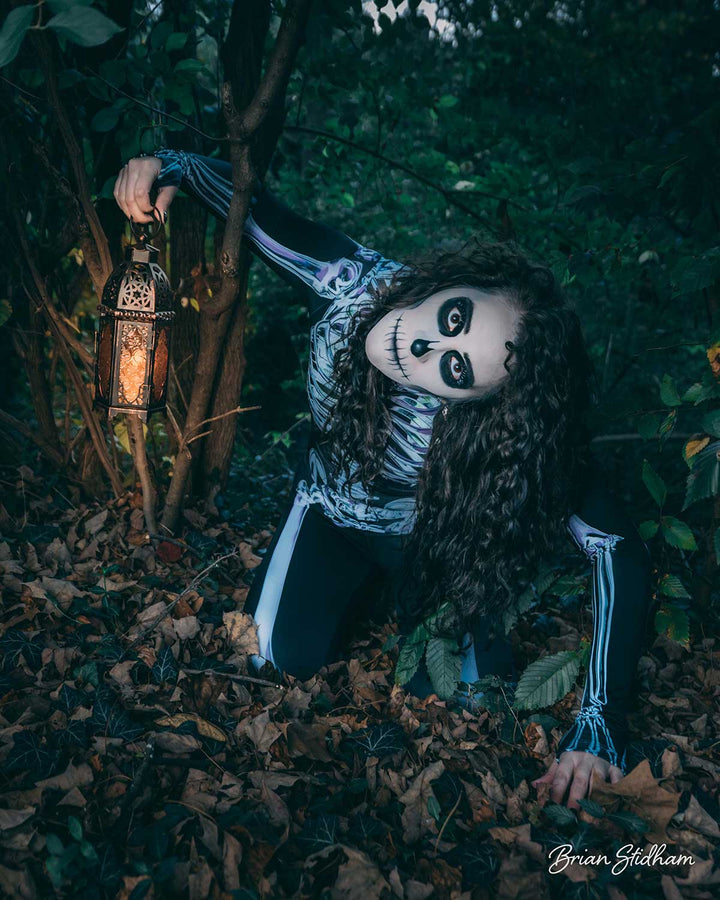 Woman wearing a black skeleton bodysuit and skull face makeup crouches in a dark forest while holding a glowing lantern.