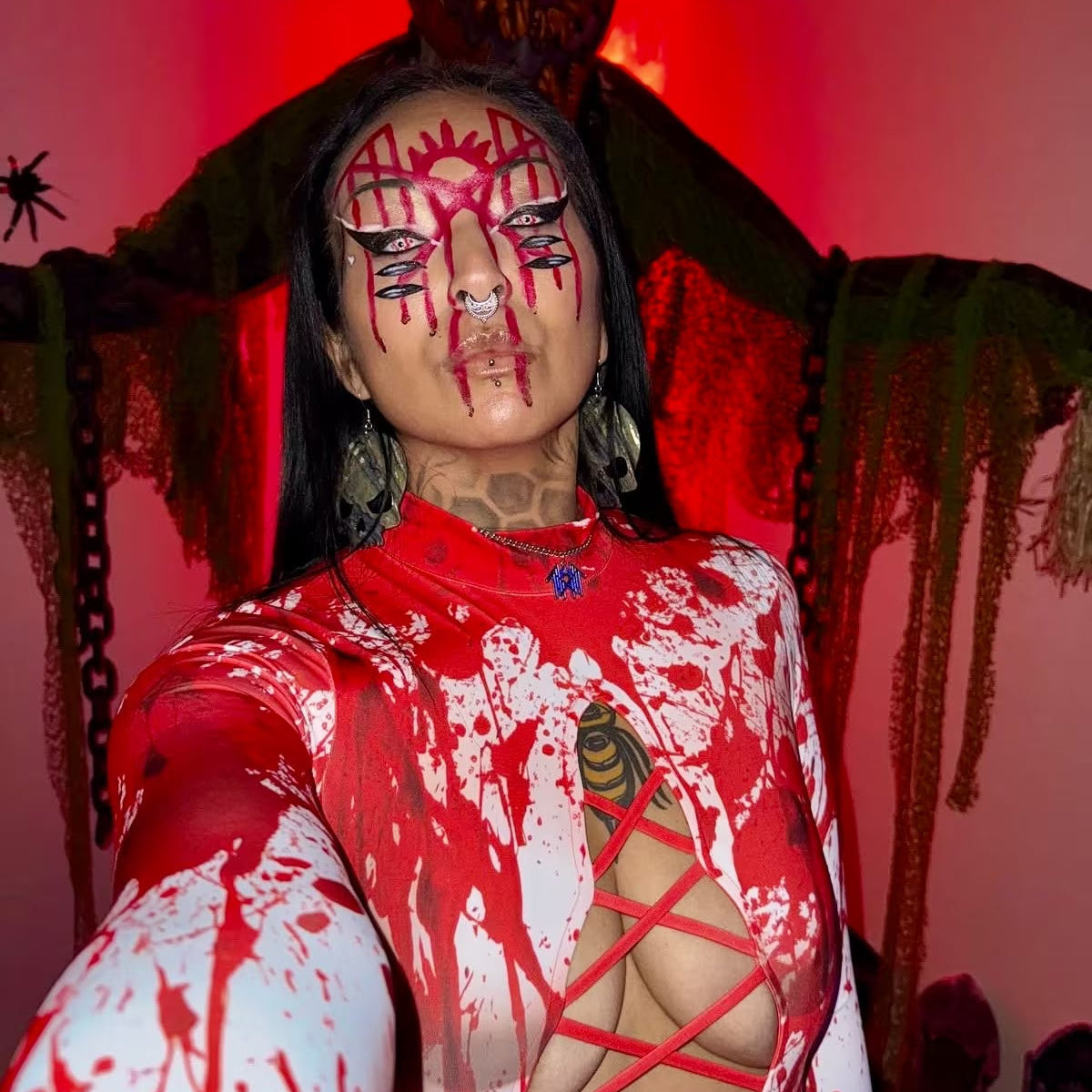 Woman wearing a red and white blood-splattered Halloween bodysuit with lace-up cutouts and dramatic red face makeup, posing in front of a creepy pumpkin-headed figure.
