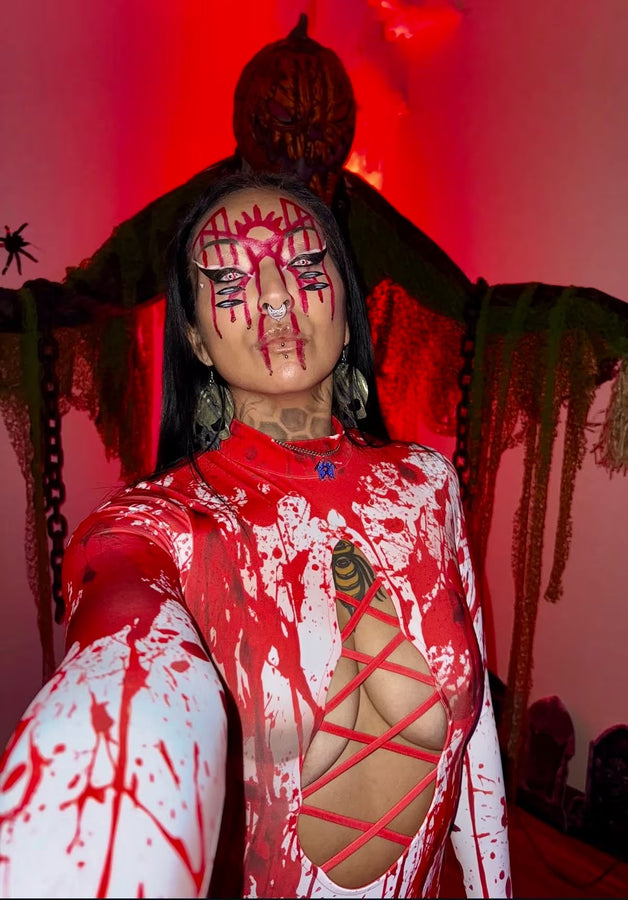 Woman wearing a red and white blood-splattered Halloween bodysuit with lace-up cutouts and dramatic red face makeup, posing in front of a creepy pumpkin-headed figure.